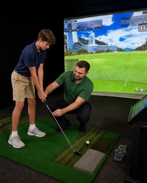 Golfer swinging a club on an indoor simulator with a large scenic screen behind him.