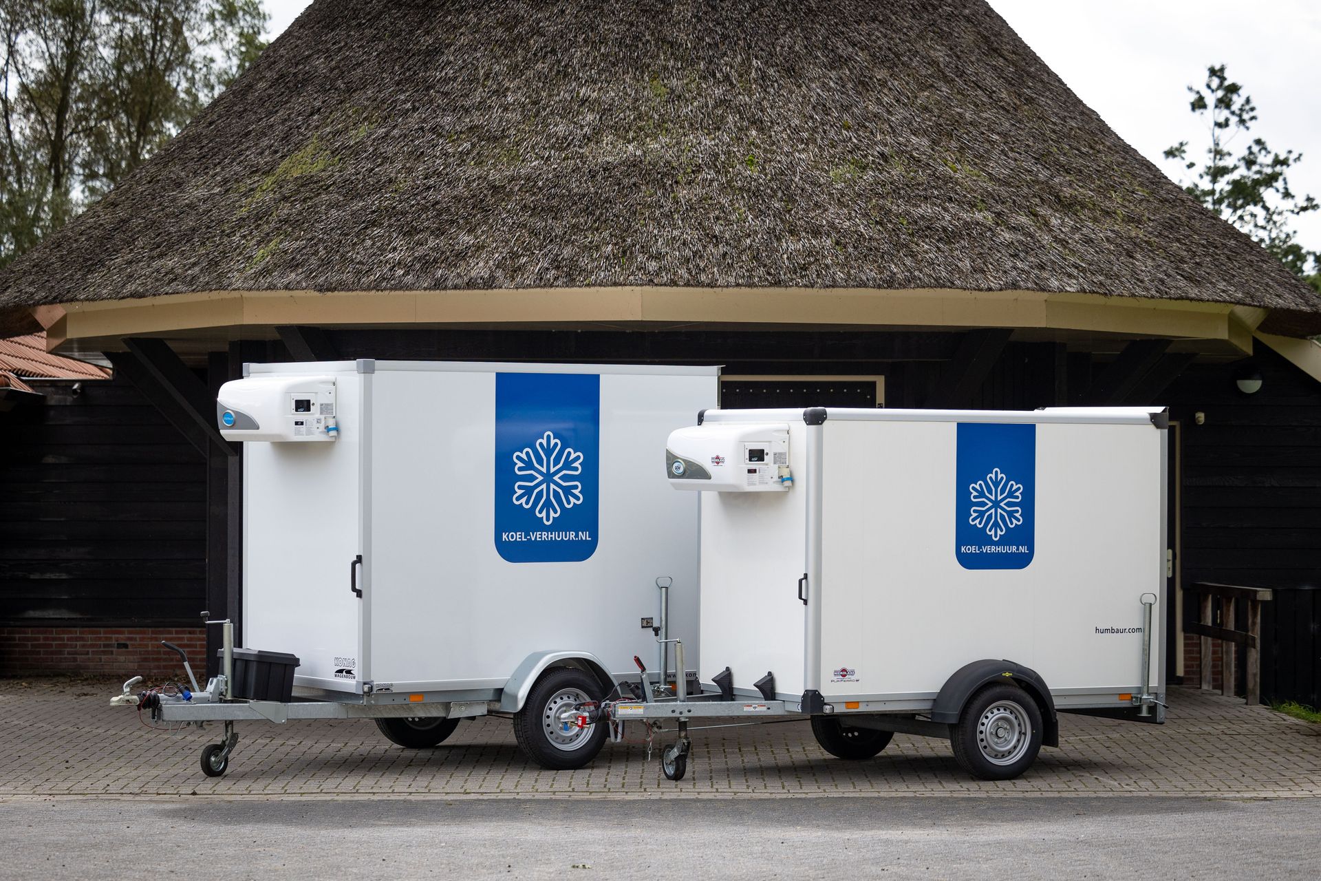 Twee witte koelwagens staan geparkeerd voor een rustiek gebouw met een rieten dak.