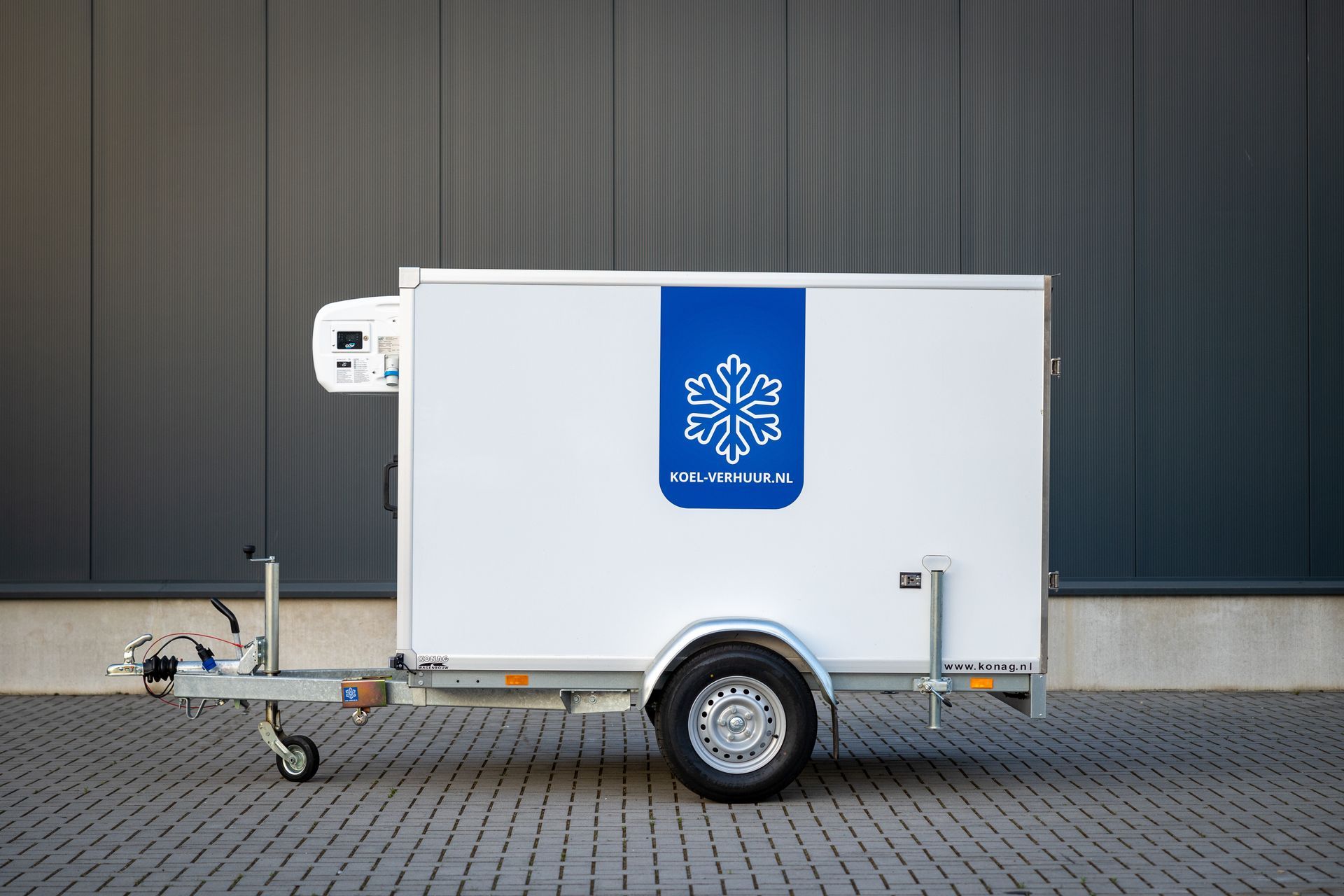 Een witte, enkelassige koeltrailer met een blauw sneeuwvloklogo staat geparkeerd op een verhard terrein tegen een donkere muur.