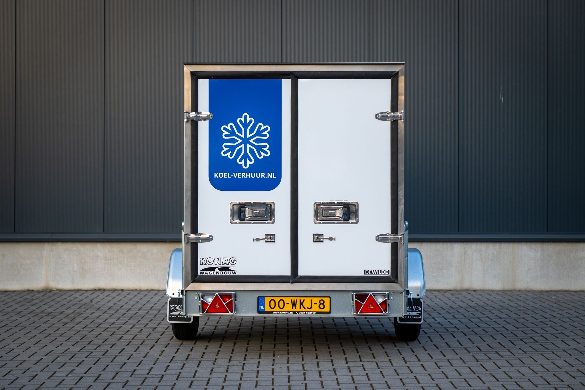 Een witte koeltrailer met een blauw sneeuwvloklogo staat geparkeerd op een grijs geplaveid oppervlak voor een golfplaten muur.