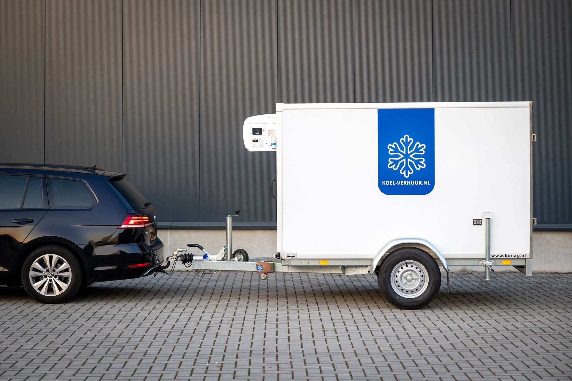 Een donkere stationwagon met een witte koeltrailer met een blauw sneeuwvloklogo staat geparkeerd op een verharde parkeerplaats.