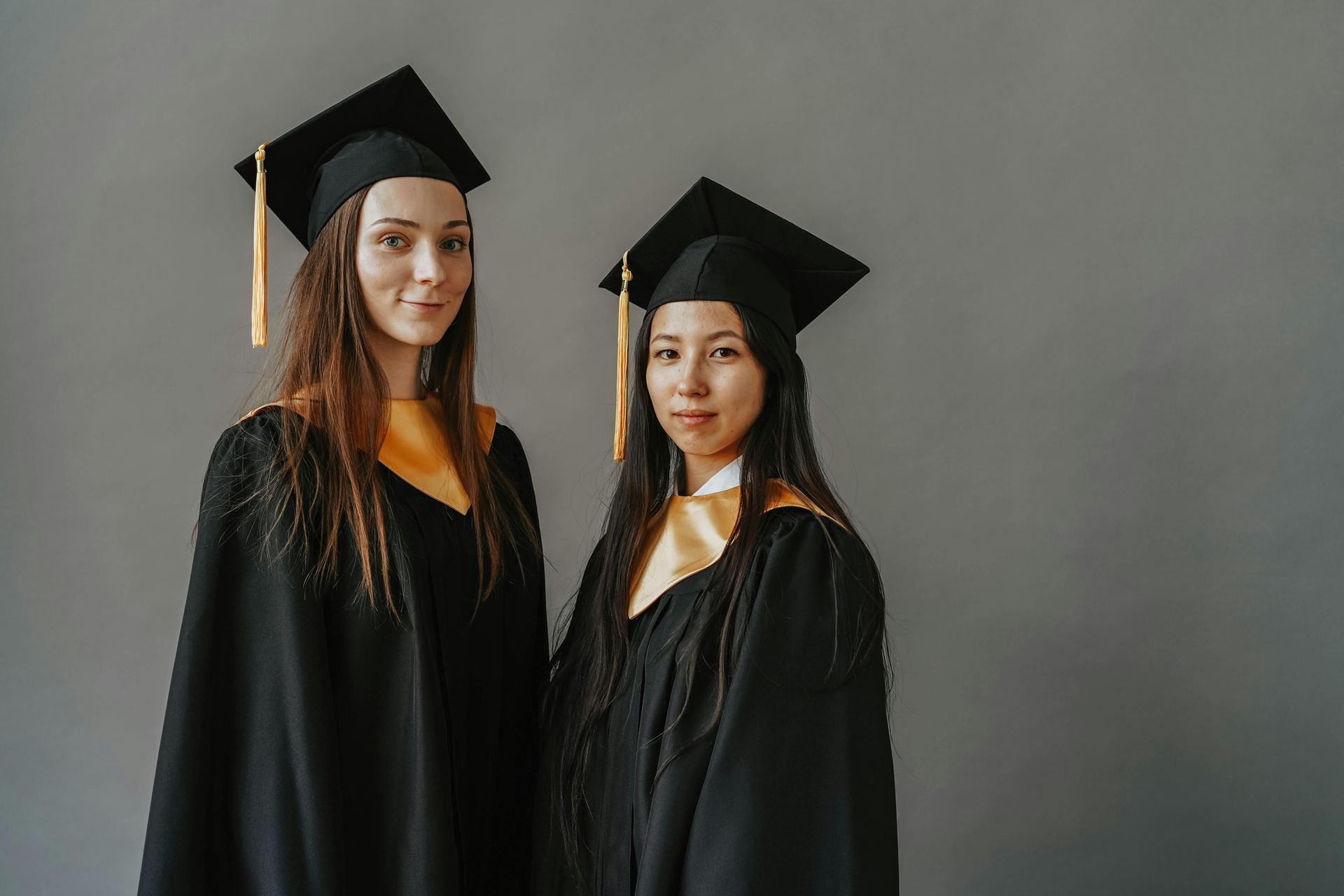 Dos graduados con togas y birretes negros con detalles dorados sobre un fondo gris.