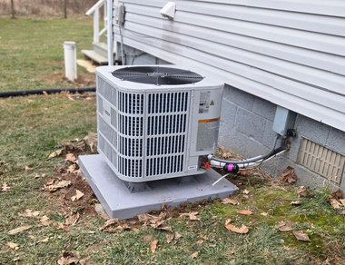 Outdoor air conditioner unit beside a house on a concrete pad, with leaves and utility lines nearby.