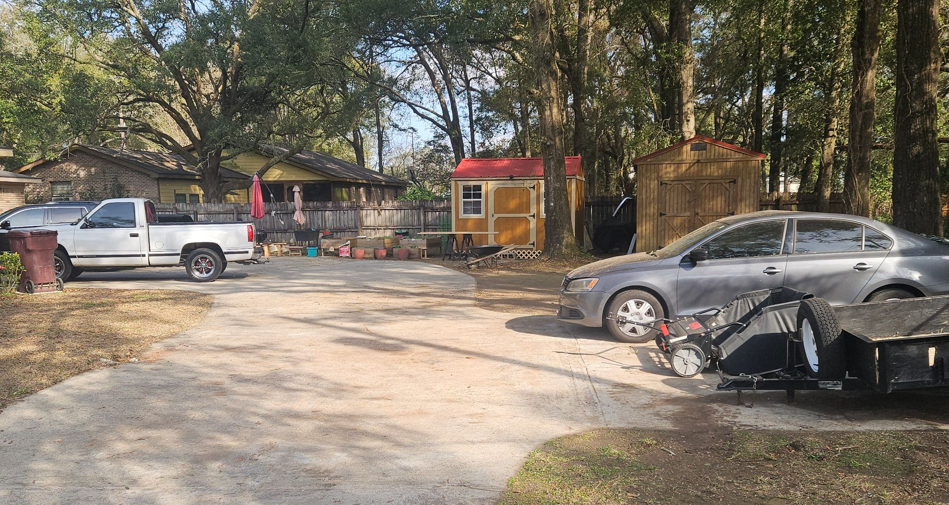 A gravel driveway with a white truck on the left, a grey sedan on the right, and two storage sheds in a wooded yard.