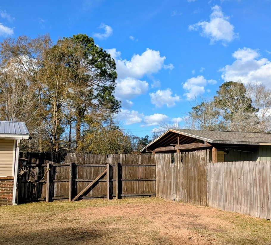 A backyard with a wooden privacy fence, a storage shed, and mature trees under a bright blue sky with scattered clouds.