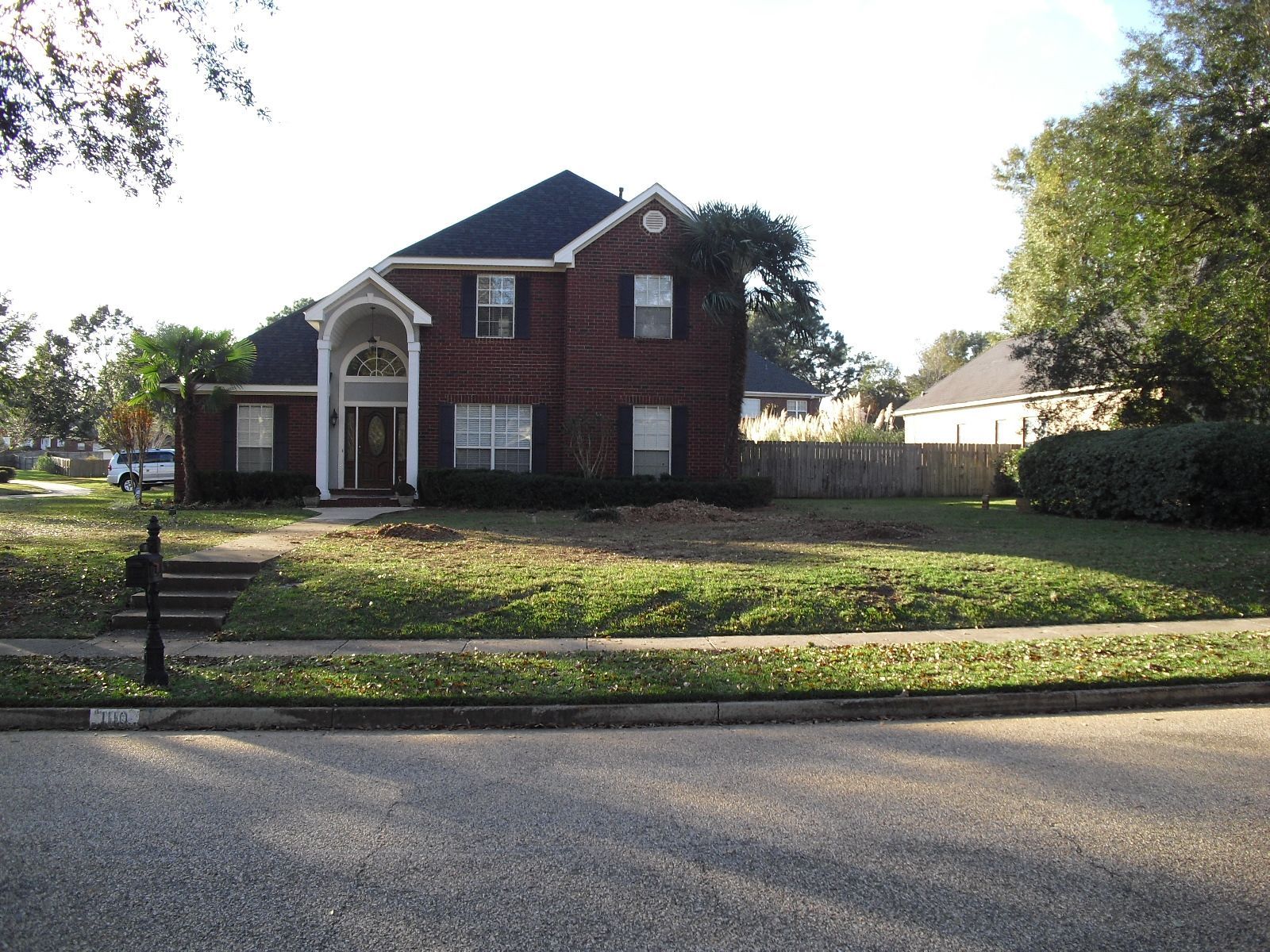 A two-story red brick house with a dark roof and white trim, situated behind a front lawn on a residential street.