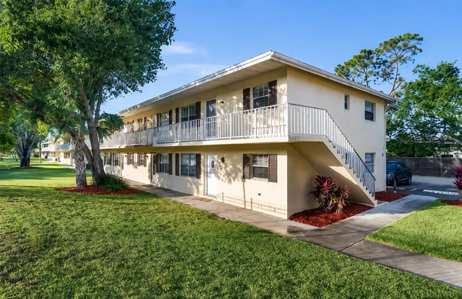Exterior of a two-story apartment building with balconies and stairs, surrounded by grass and trees.