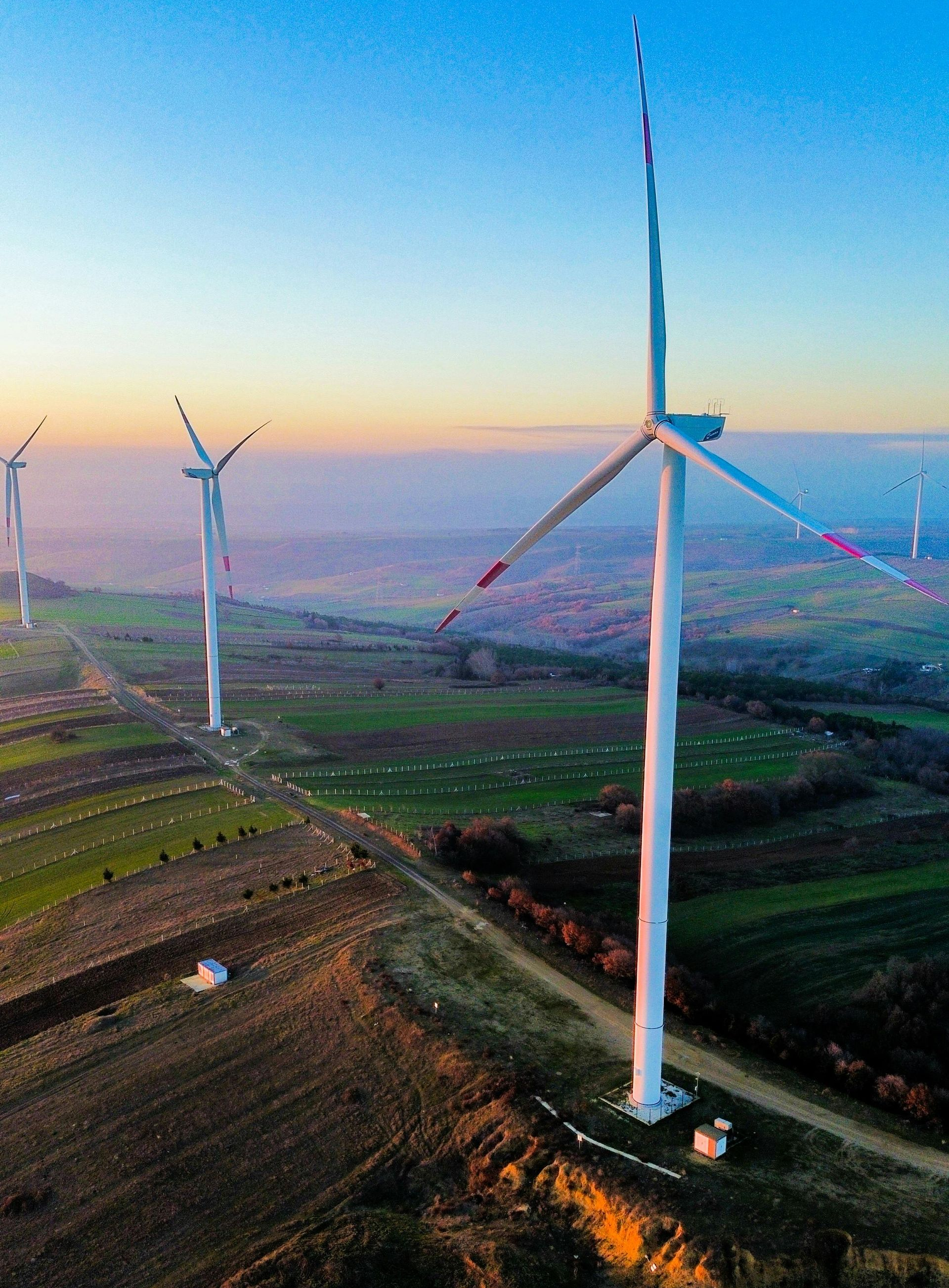 Wind turbines standing on a green hillside at sunset, with fields and a hazy sky.