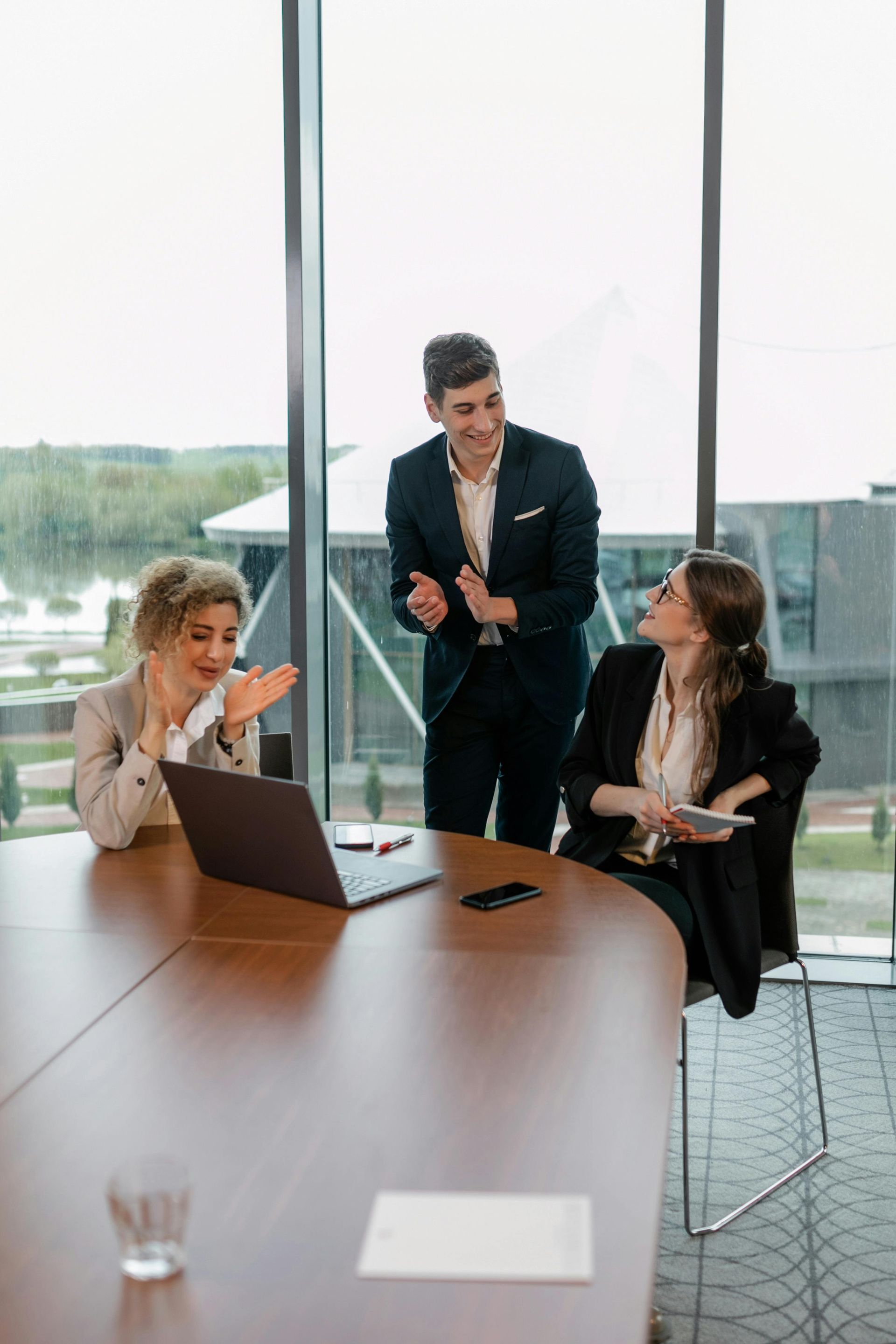 Three colleagues in a meeting around a laptop and notepad by a large windowed conference room