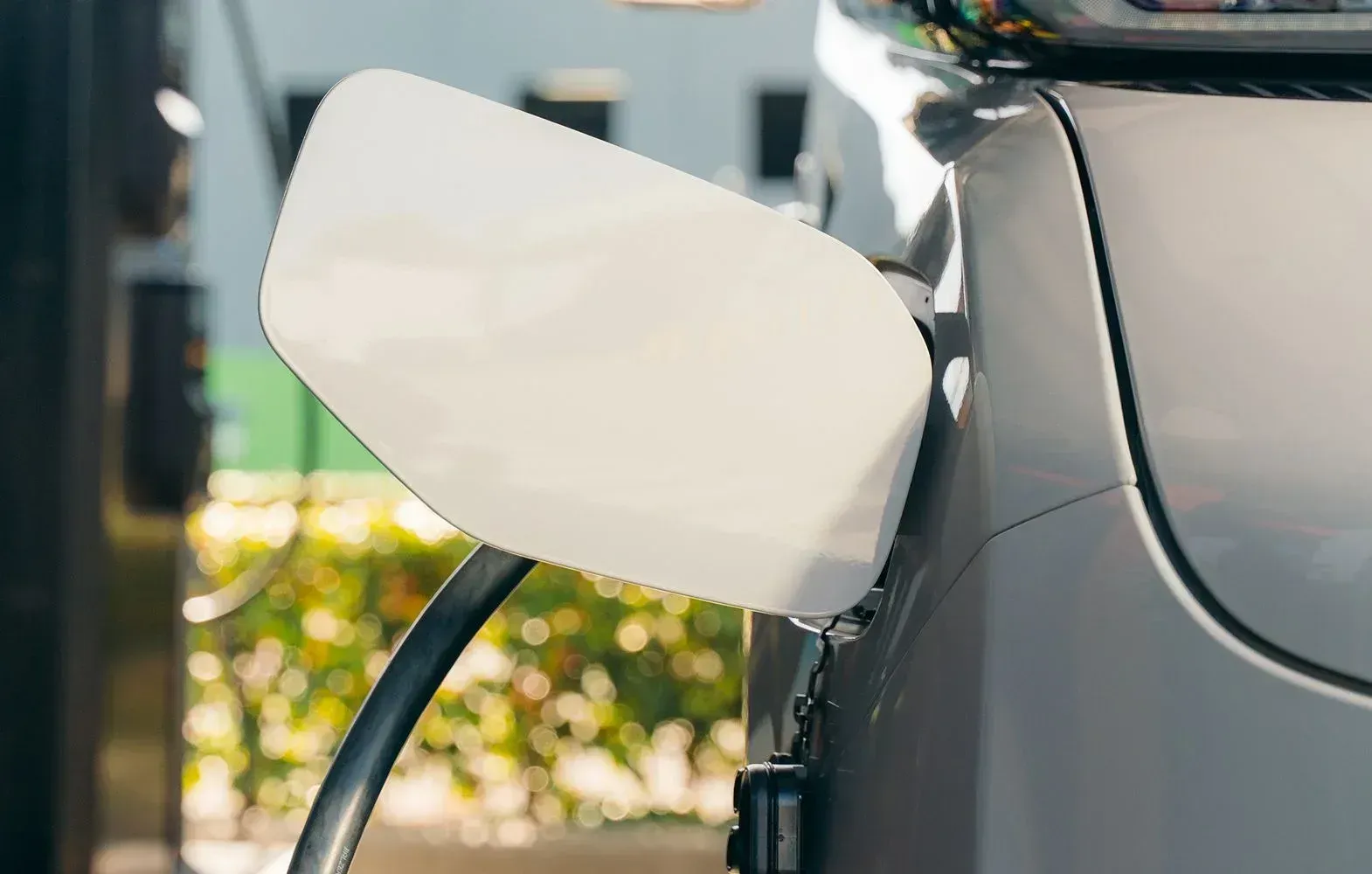 A charging cable plugged into the side of a silver electric vehicle, outdoors in front of out-of-focus green foliage.
