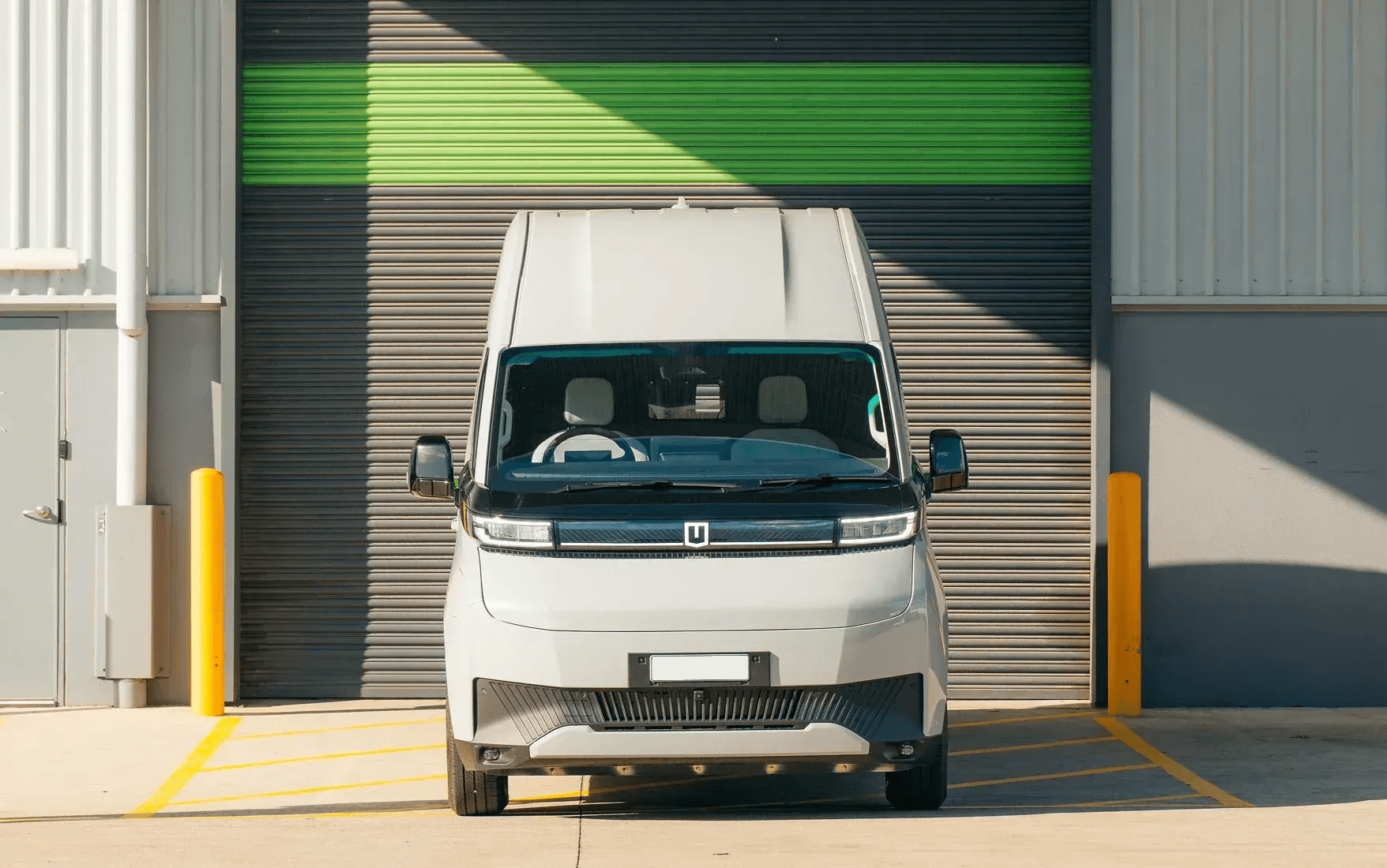 A light gray box van parked facing forward in front of a closed industrial warehouse door with a green horizontal stripe.