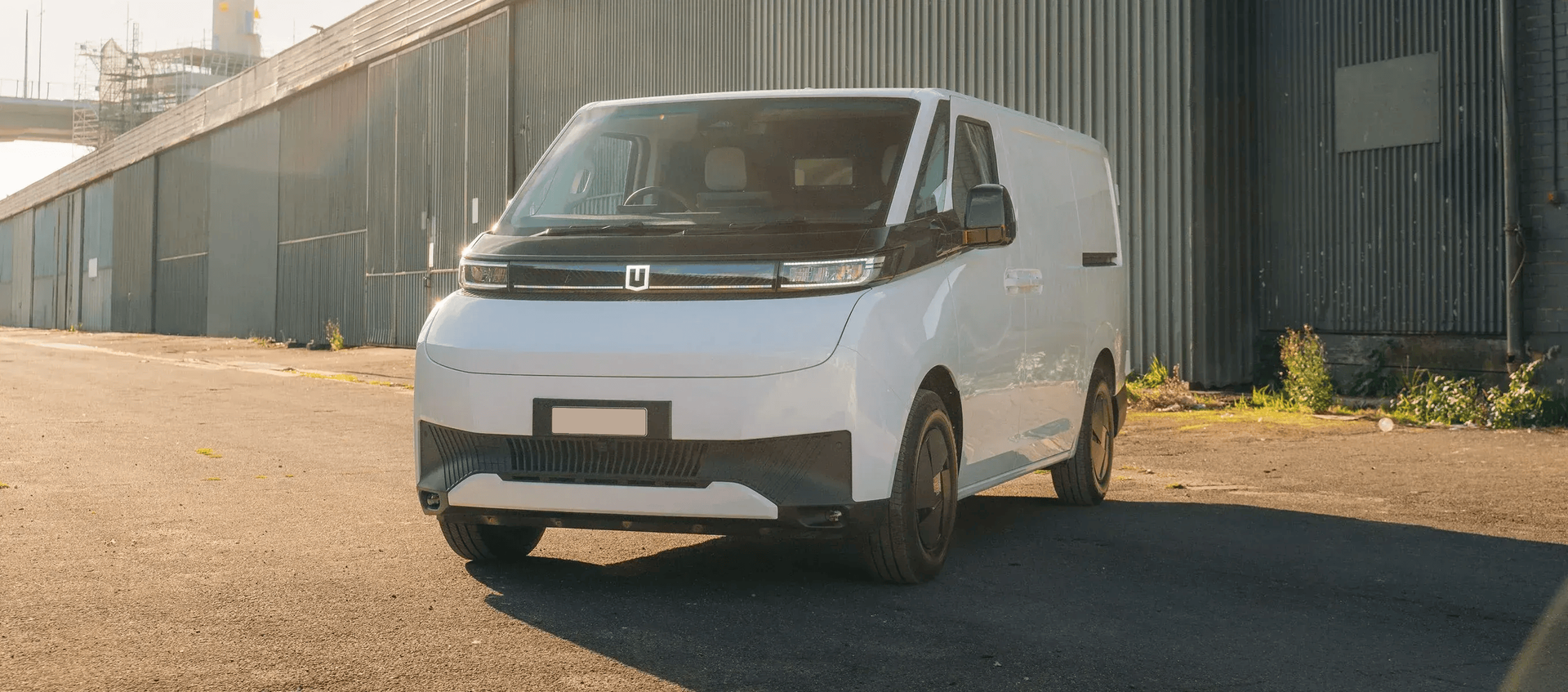 A white electric cargo van parked on a gravel lot near a large industrial building at sunset.