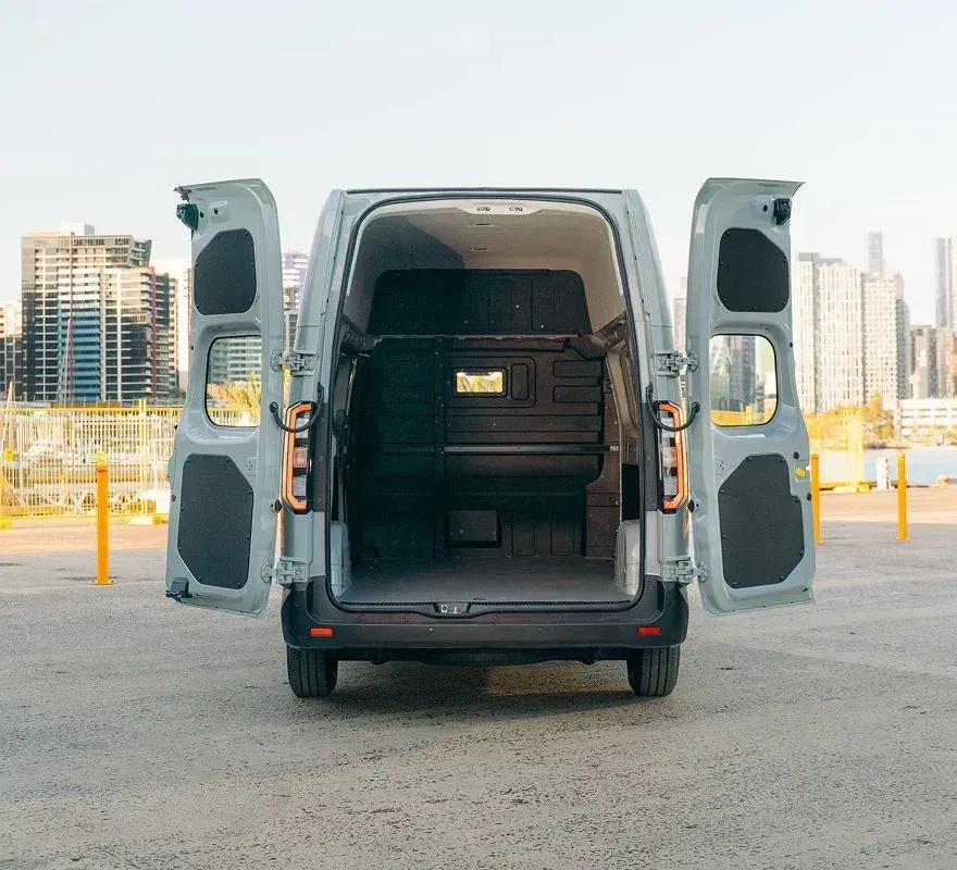 Rear view of a light gray commercial cargo van with its double back doors open, parked against a city skyline background.
