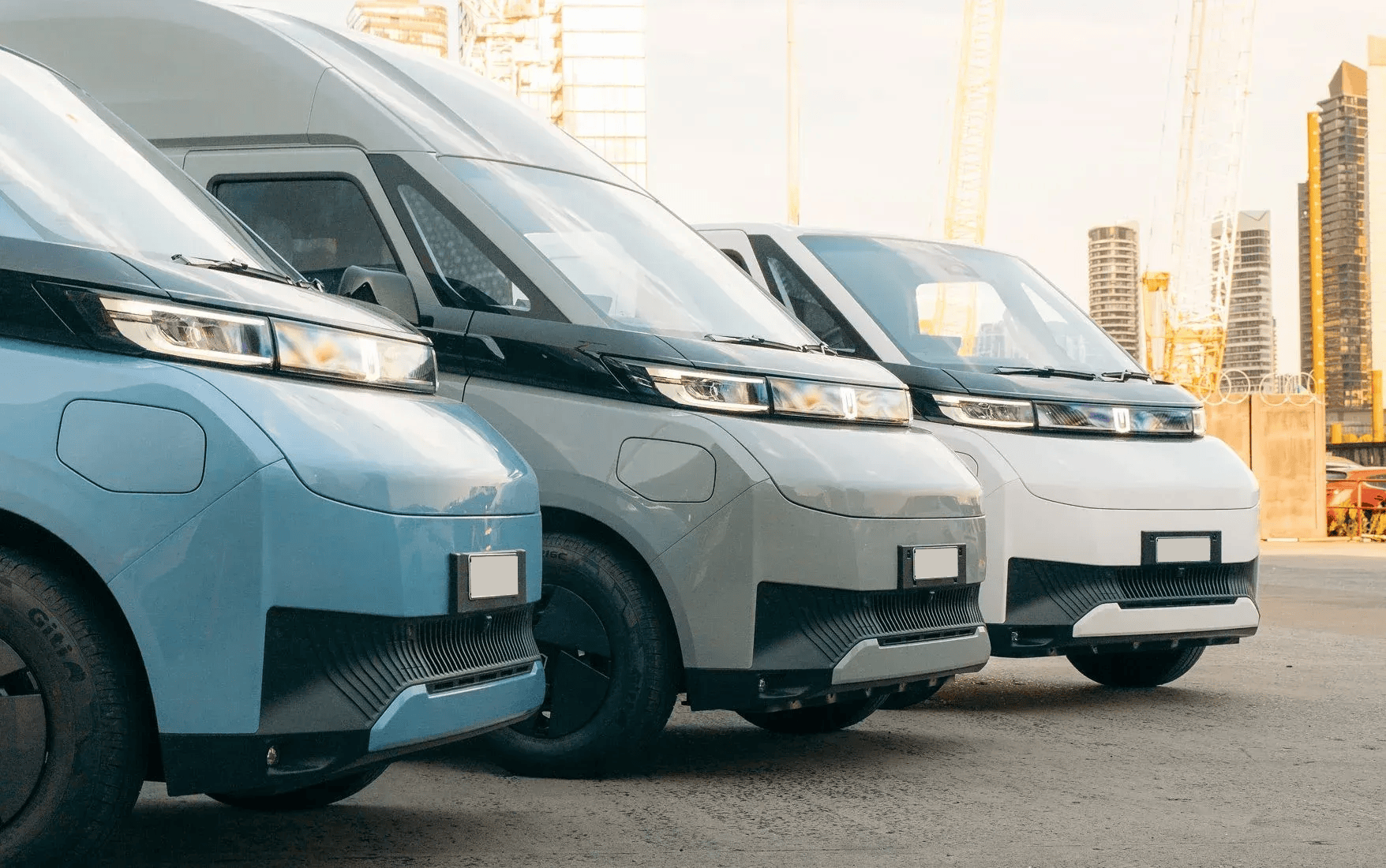 Three parked electric vans in blue, grey, and white, positioned in a line against an urban background.