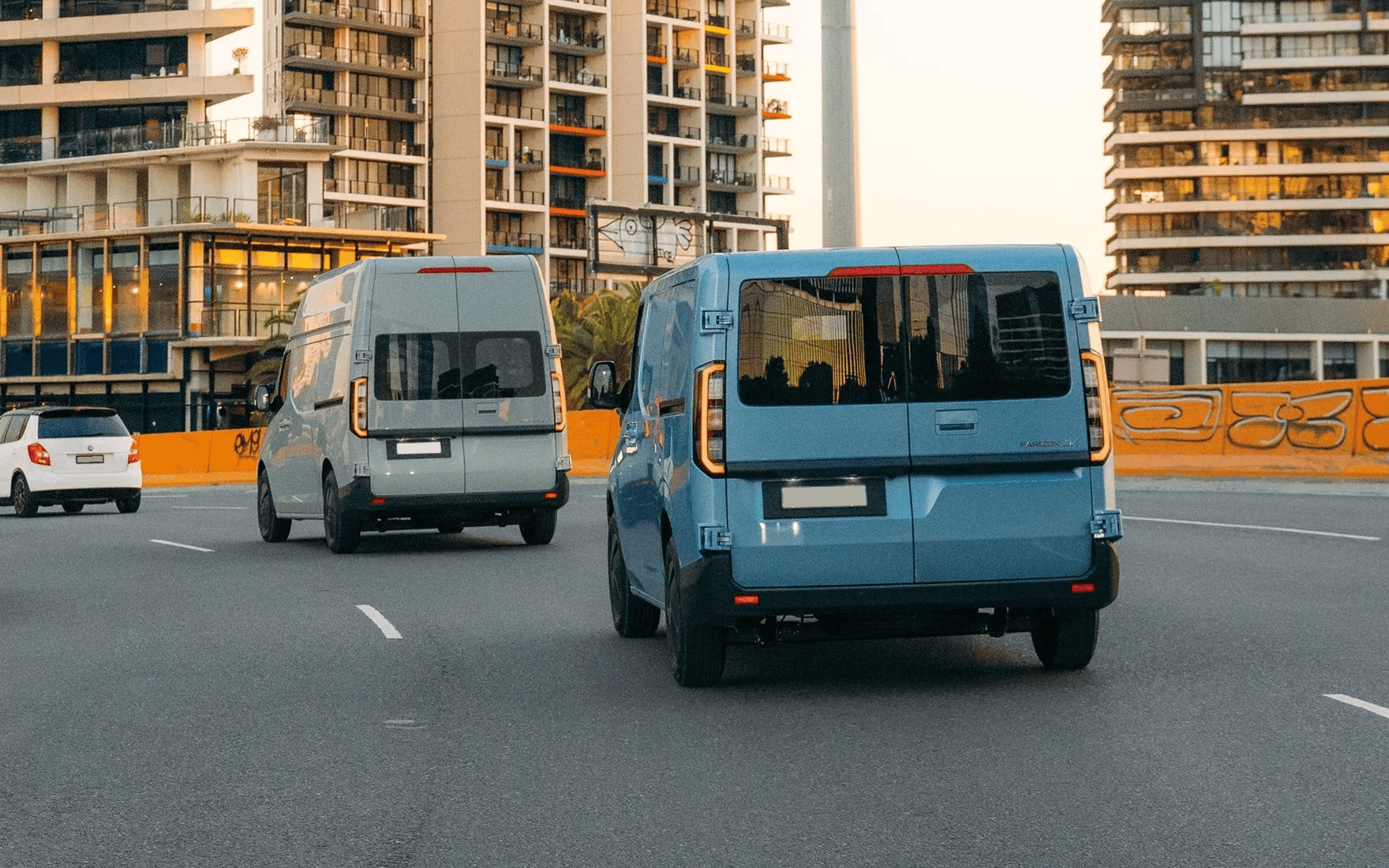 A light blue van and a gray van driving on a paved road in a city with apartment buildings in the background.