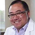 Smiling person in glasses and a white coat, seated indoors near a window.