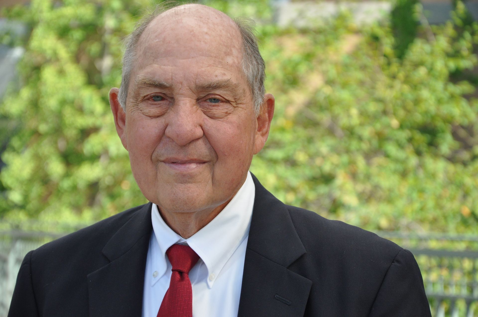 Smiling man in a black suit, white shirt, and red tie standing outdoors with greenery behind him