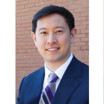 Portrait of a smiling person in a navy suit and purple striped tie against a brick wall