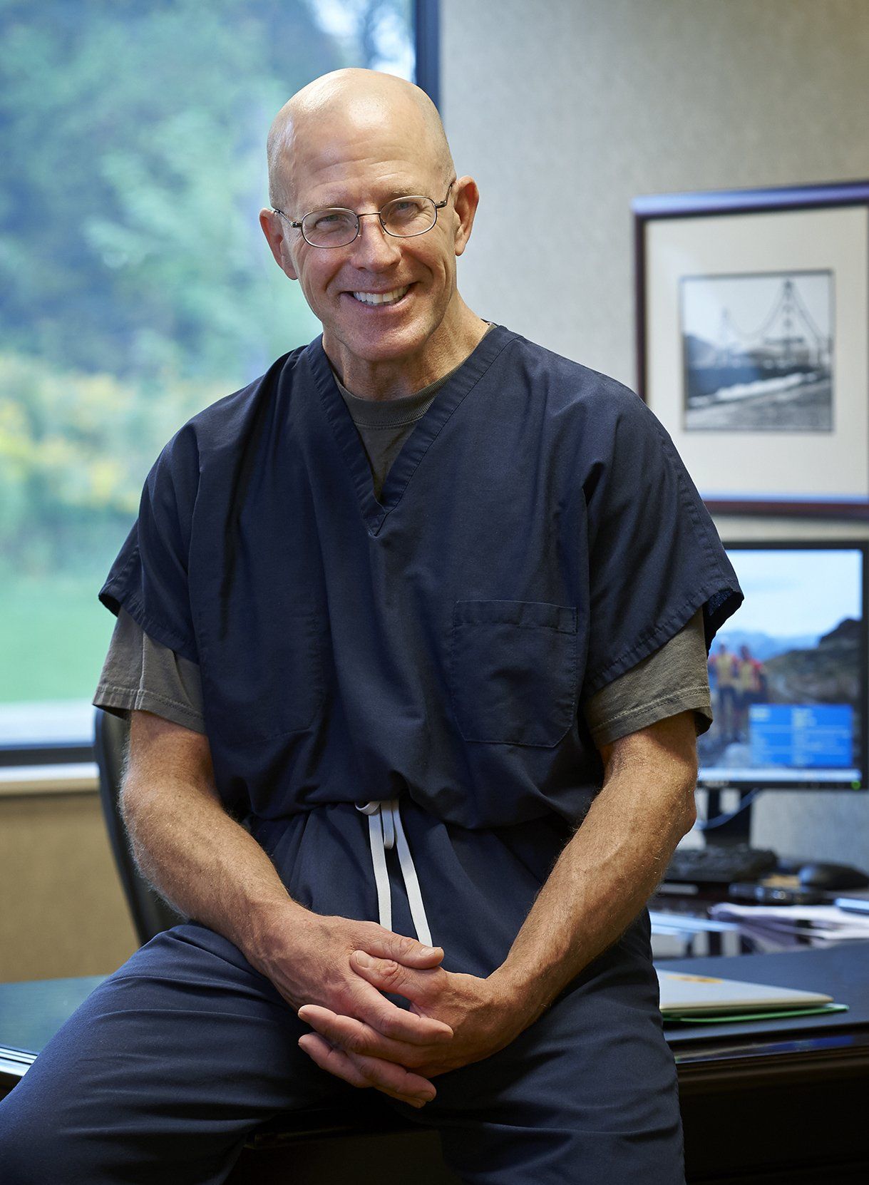 Smiling bald man in dark scrubs standing in an office, with framed pictures on the wall.