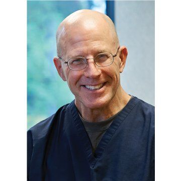 Smiling bald man wearing glasses and dark blue scrubs against a soft blue-green background
