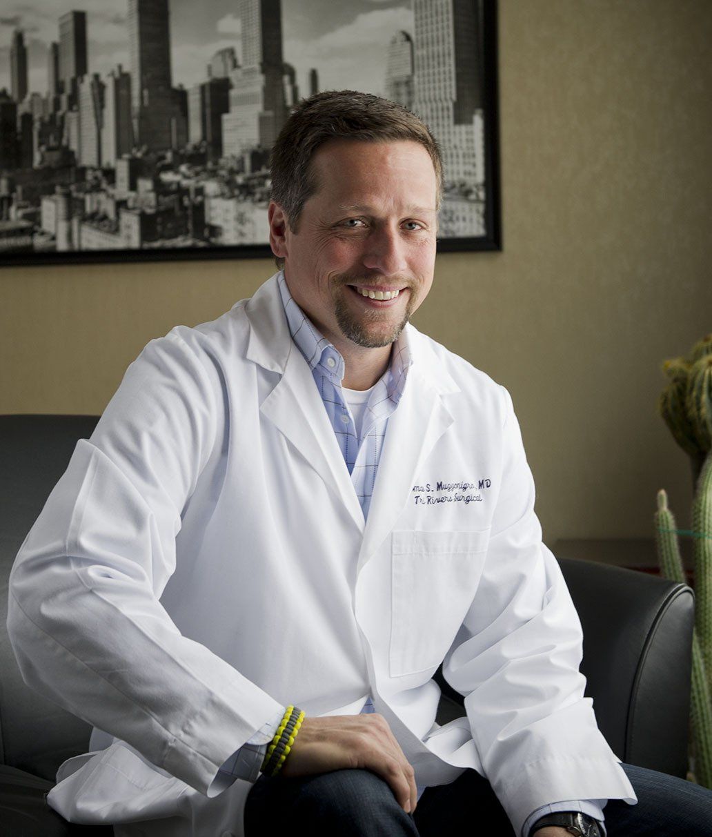 Smiling doctor in a white coat seated in an office, with a city skyline photo behind him.