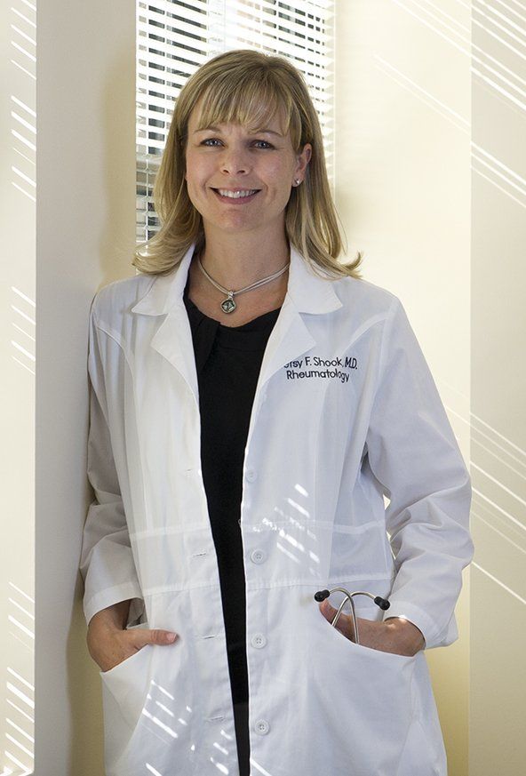 Woman in a white lab coat standing indoors by a window with blinds, hands in pockets, smiling