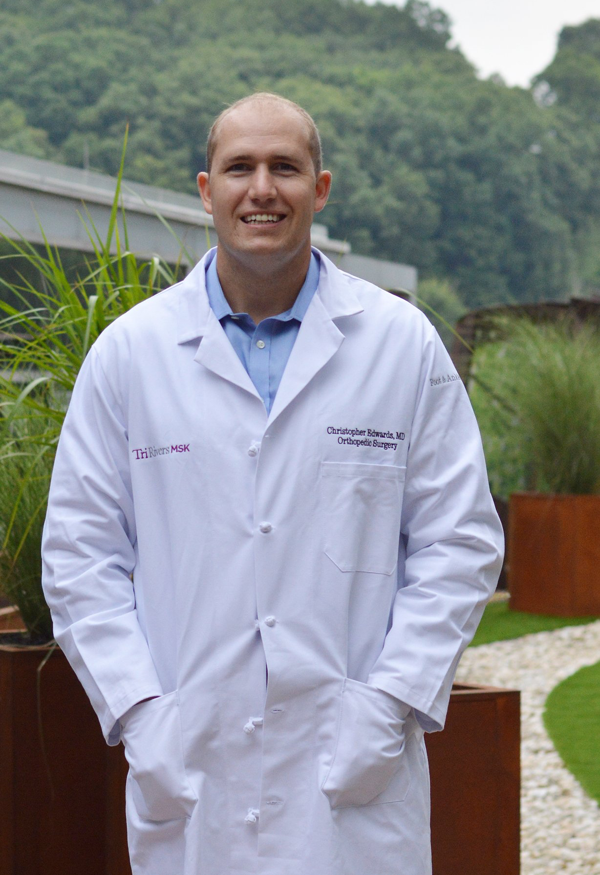 Man in a white lab coat standing outdoors in front of greenery and a building