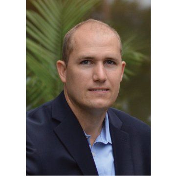 Portrait of a man in a dark blazer and light blue shirt, standing outdoors with blurred greenery behind him
