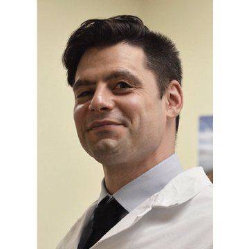 Smiling man in a white lab coat and black tie indoors, shown from the shoulders up.