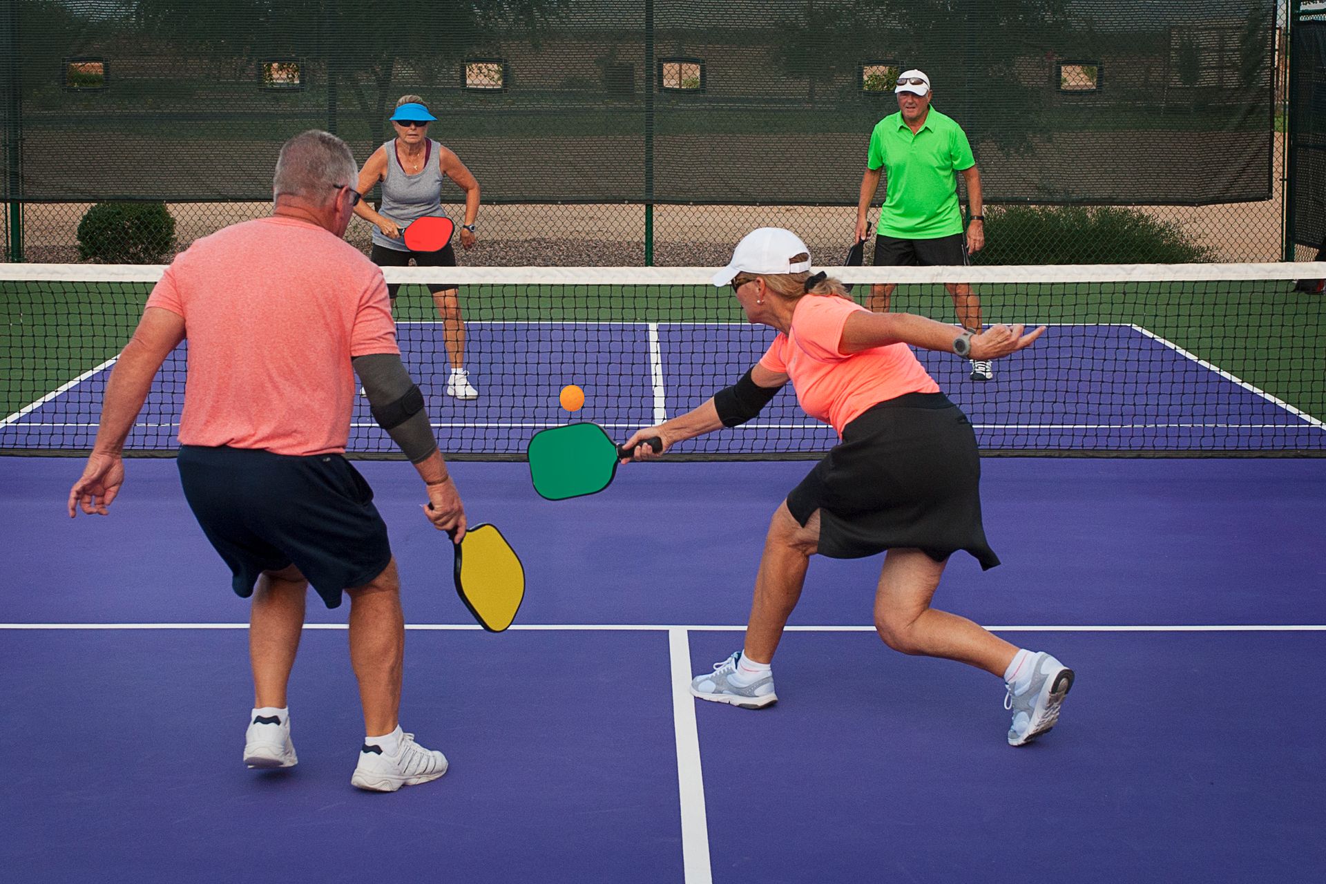 Players in a pickleball game on a blue court, two in the foreground reaching for the ball near the net.