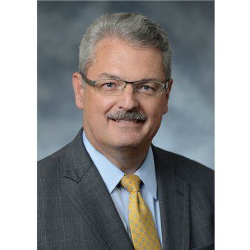 Portrait of a smiling man in a gray suit, light blue shirt, and yellow tie against a gray background