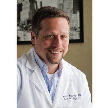 Smiling man in a white lab coat standing in an office with framed wall art