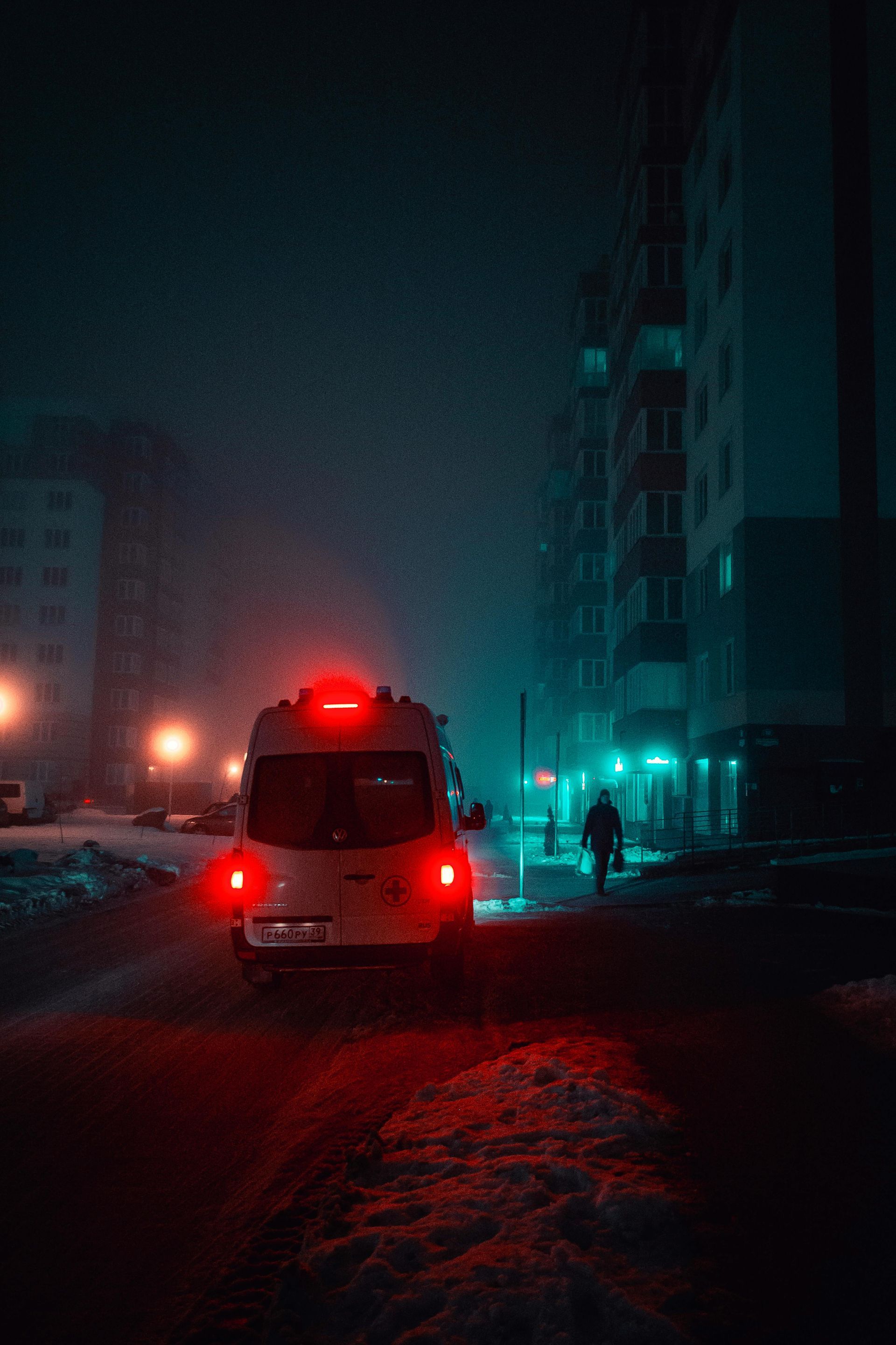 Red emergency vehicle on a snowy city street at night, with glowing lights and tall buildings in fog.