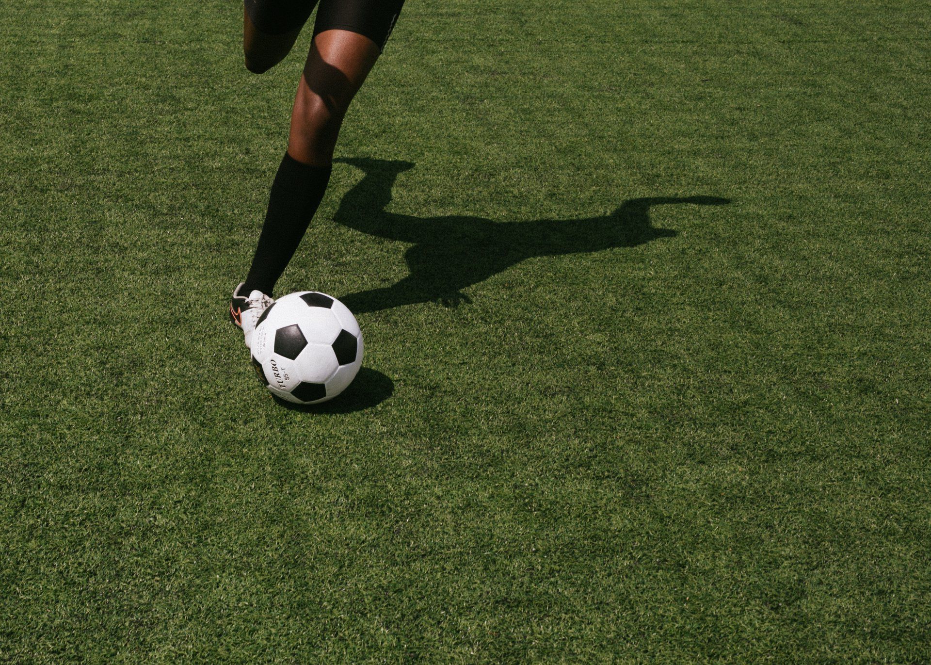 Soccer player’s leg in black socks kicking a black-and-white ball on green grass