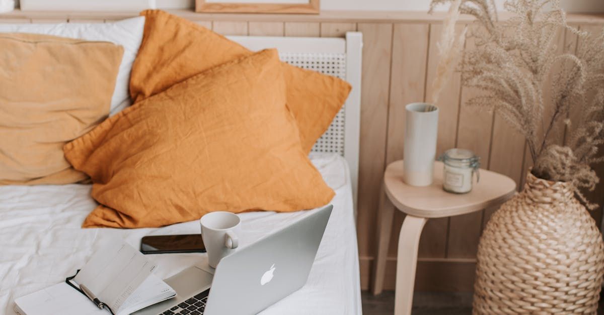 Cozy bed with orange pillows, open laptop, and a small round side table beside a woven basket lamp