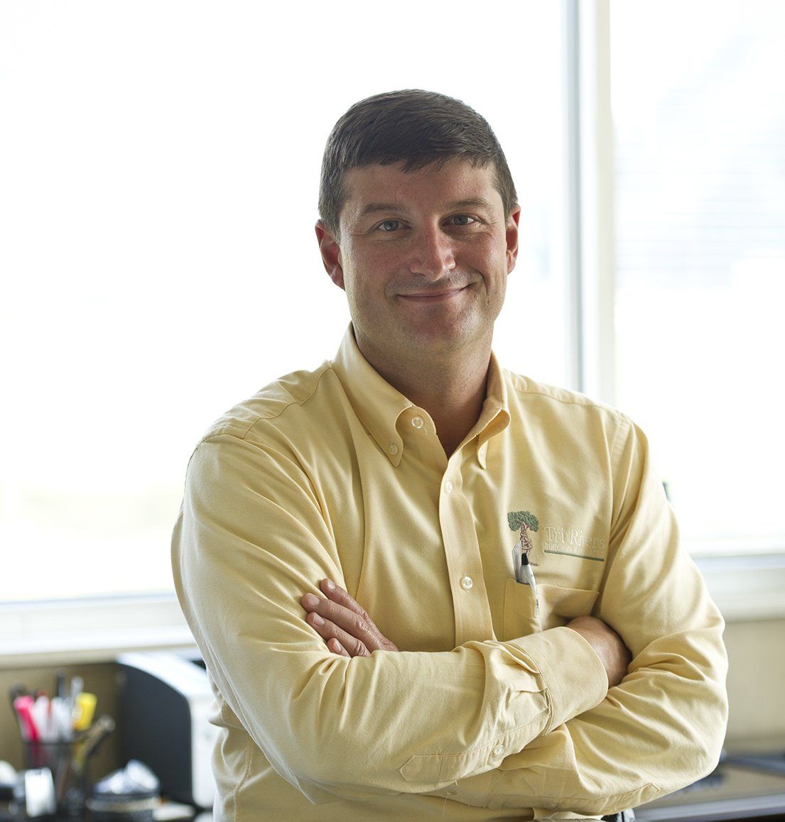 Man standing with arms crossed in a bright office, wearing a light yellow shirt and smiling.