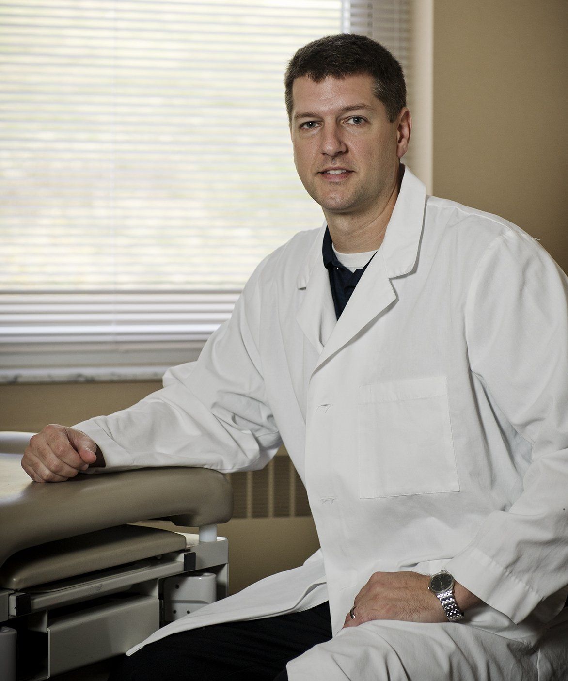 Man in a white lab coat sitting beside an exam table in a medical office