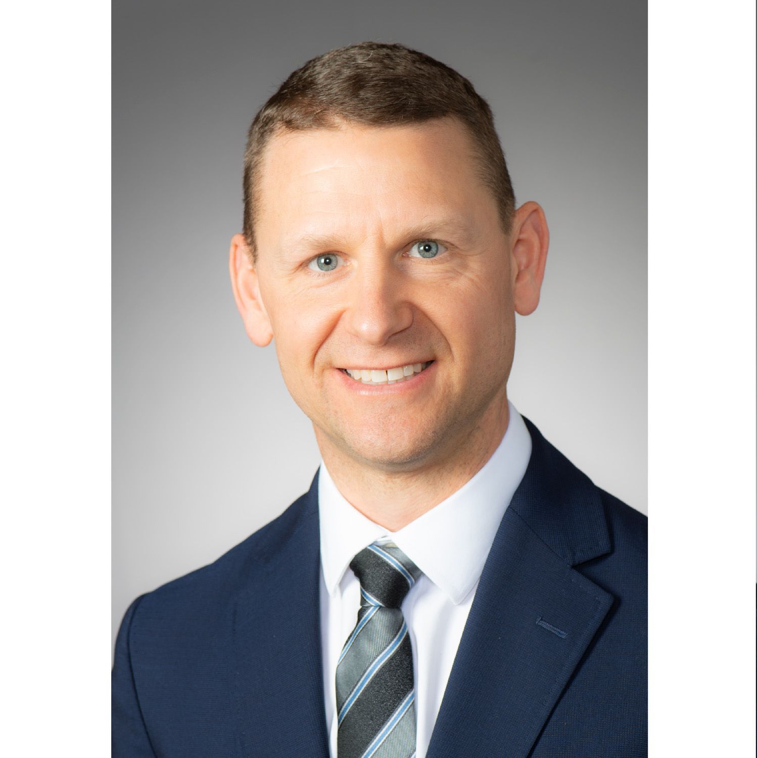 Professional headshot of a smiling person in a navy suit and striped tie against a gray background