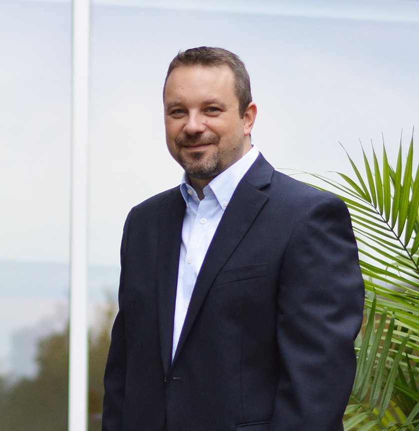 Man in a navy suit standing outdoors beside a palm plant, smiling at the camera.