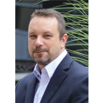 Man in a navy blazer and light shirt posing outdoors near green plants