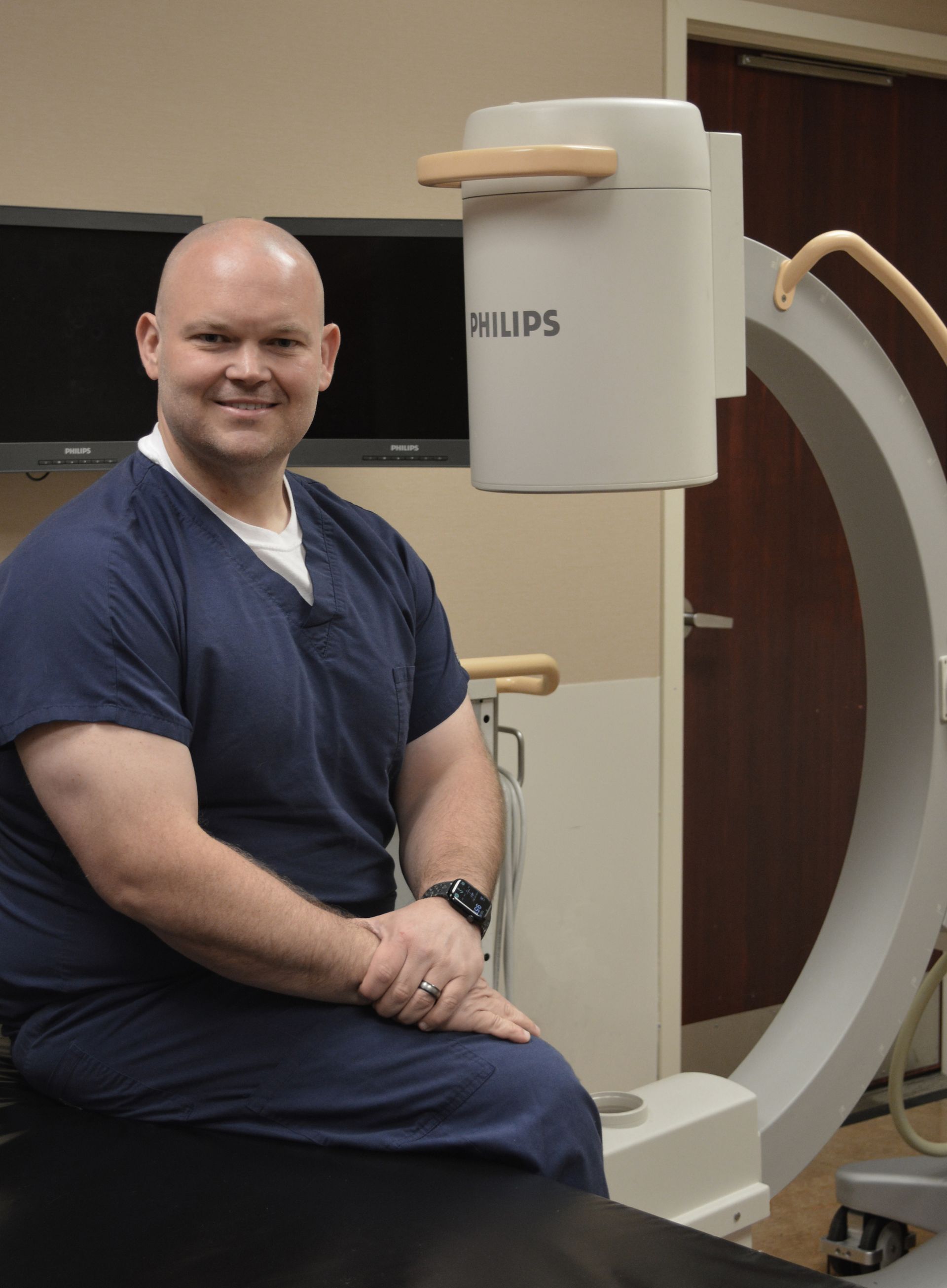 Smiling clinician in navy scrubs seated beside a Philips medical imaging machine