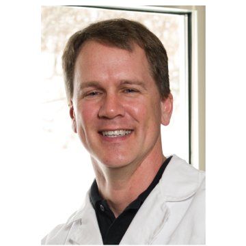 Smiling man in a white lab coat, standing indoors near a bright window.
