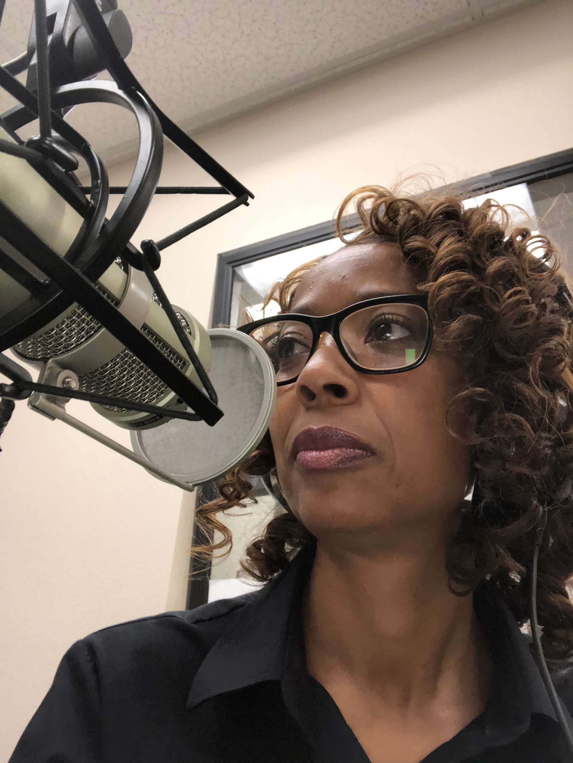 Woman speaking into a microphone in a recording studio, wearing glasses and a black shirt.