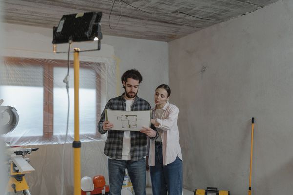 Due persone esaminano i progetti in una stanza non ancora ultimata, illuminata da lampade da cantiere e dotata di attrezzi.