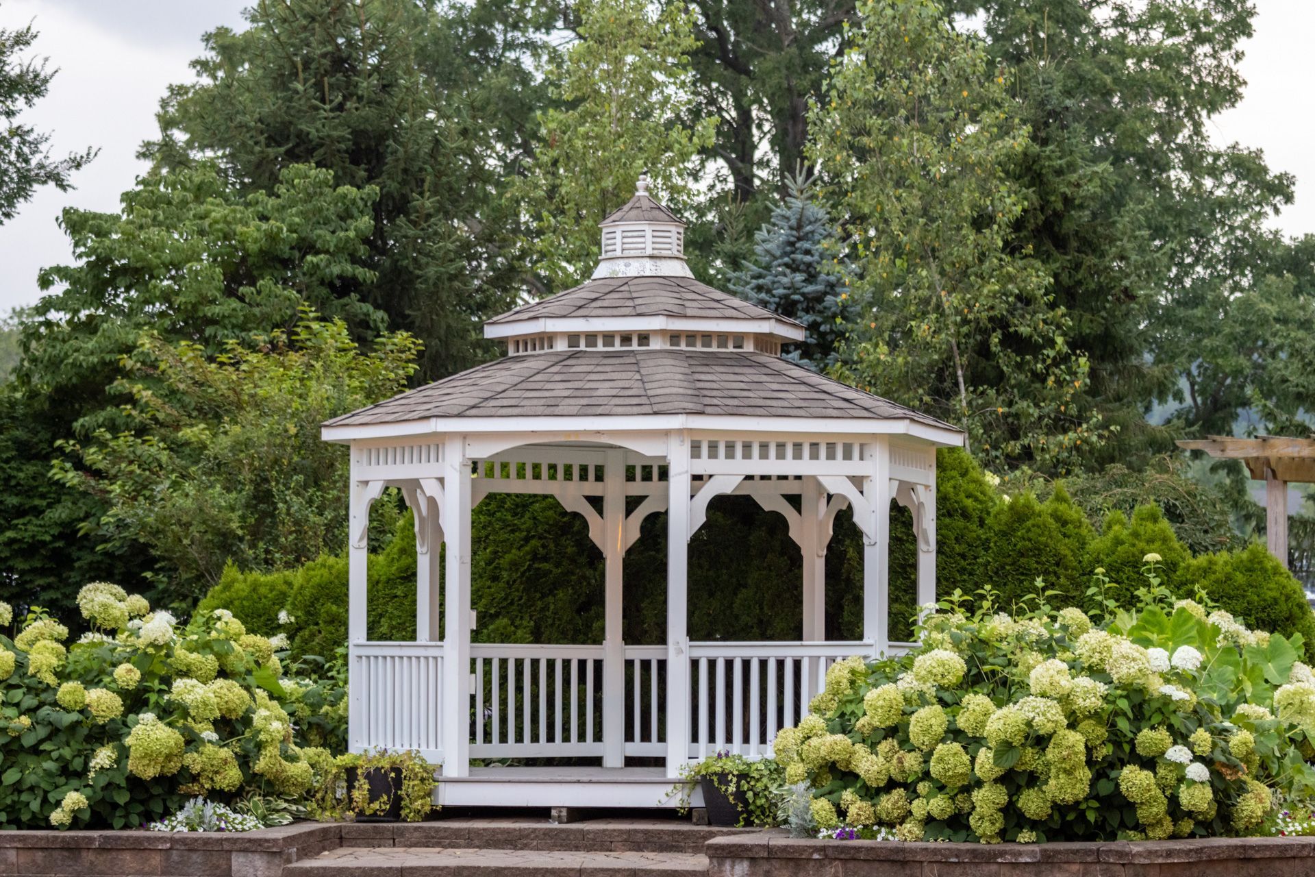 A white gazebo is surrounded by trees and flowers in a garden.