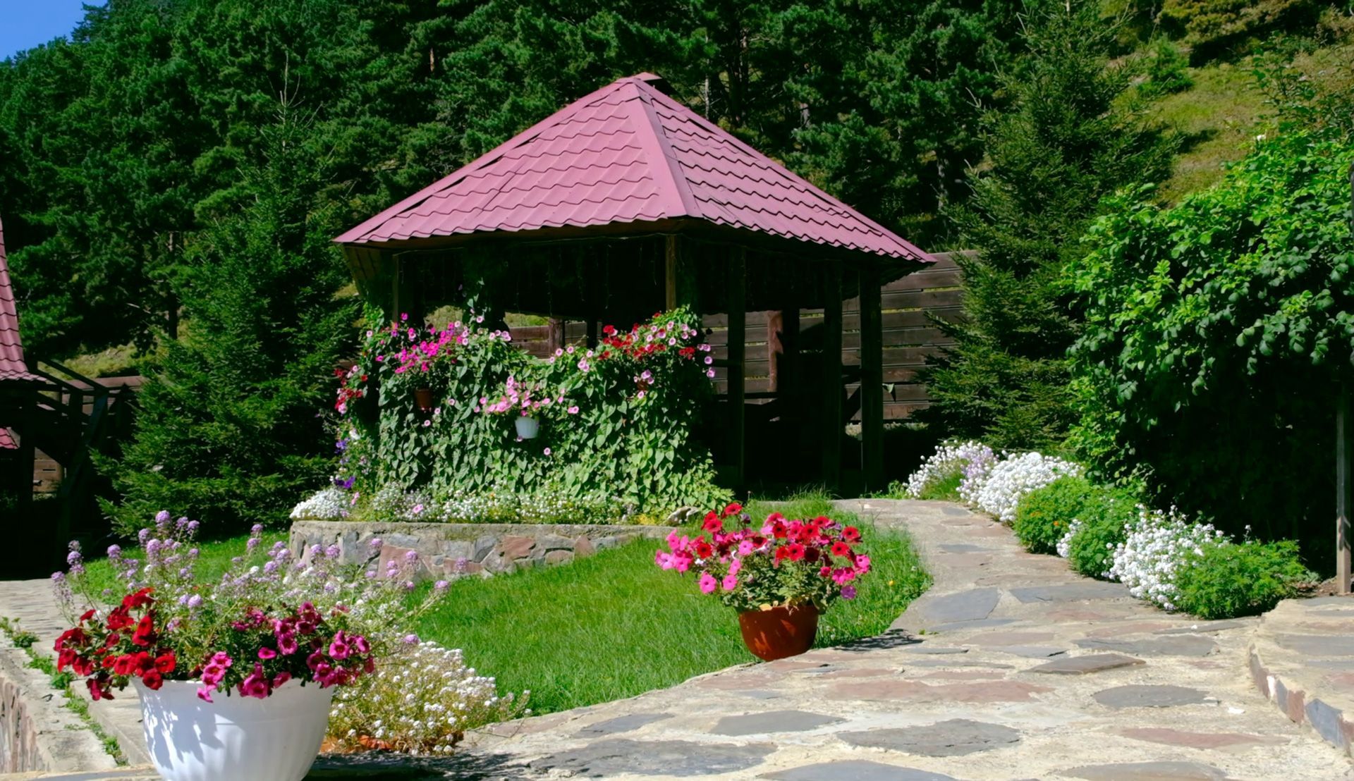 A gazebo with a red roof is surrounded by flowers and trees.