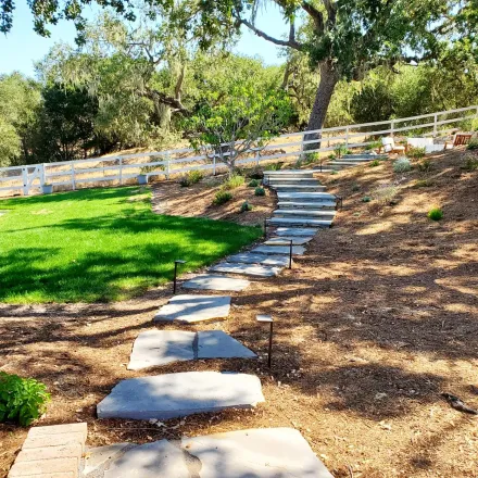 A stone walkway leading up to a grassy hill with a fence in the background.