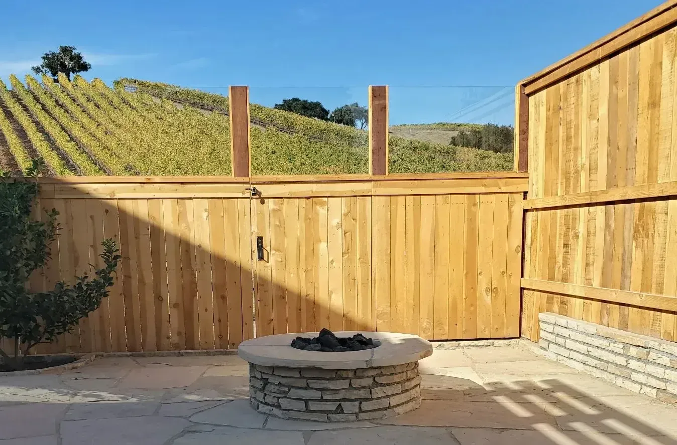 A fire pit in front of a wooden fence with a vineyard in the background.