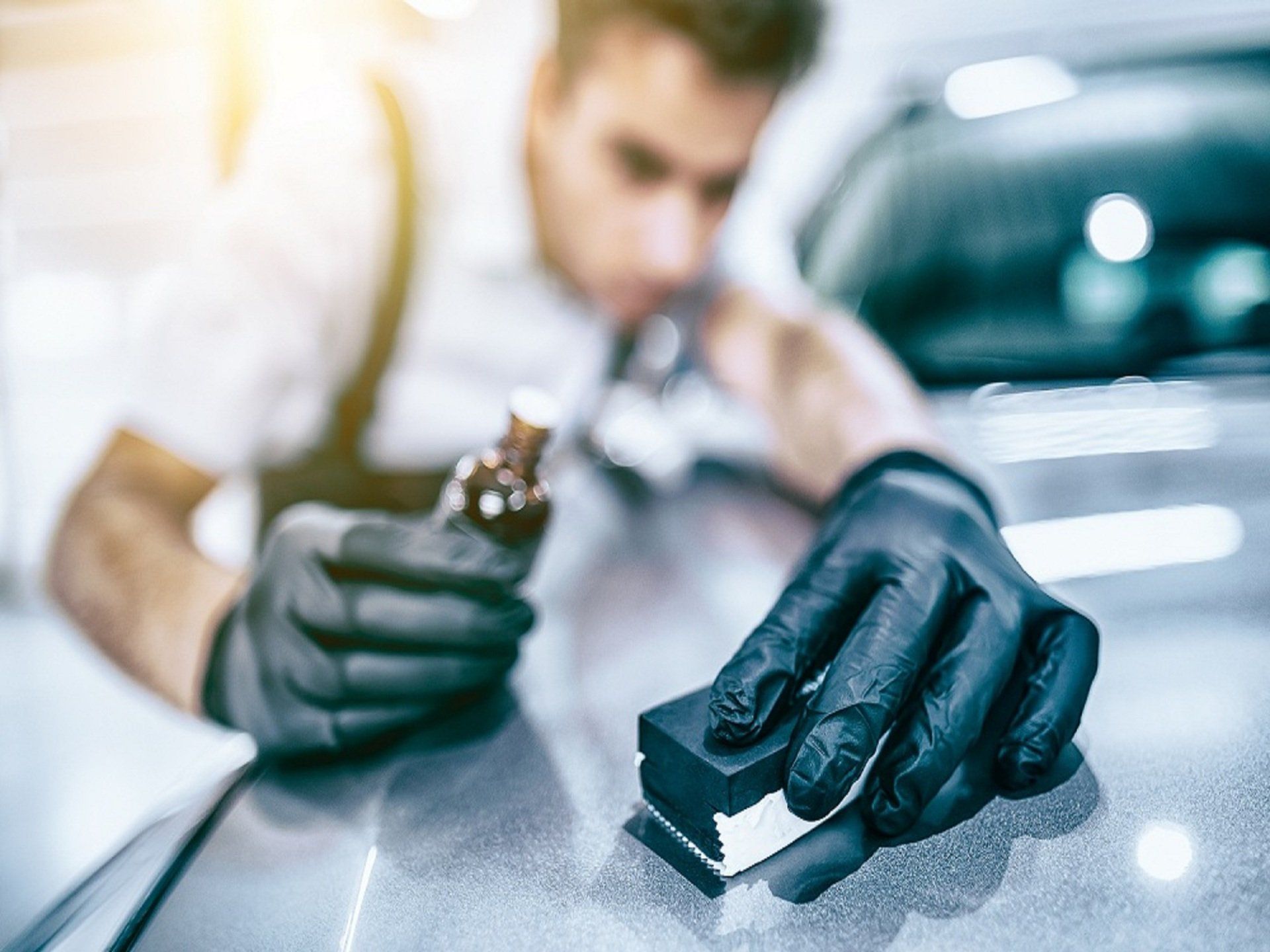 Gloved mechanic polishing a silver car with a black sponge while holding a polish bottle