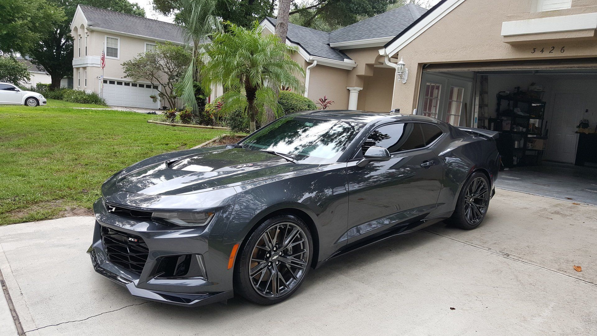 Dark gray sports car parked in a driveway beside an open garage in a suburban home.